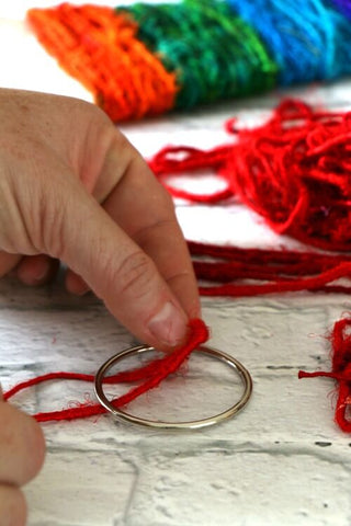 Woman's hands threading red yarn around a metal ring next to yarn sample cards on a white surface