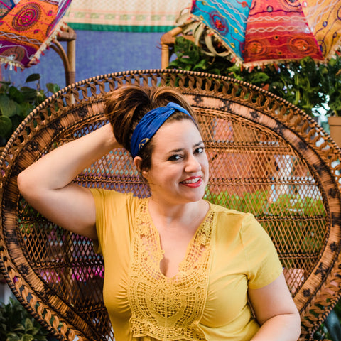 A plus sized woman wearing a yellow shirt is touching her hair. In her brown hair is a blue kameela knot headband, made out of reclaimed sari silk