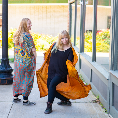 Two women wearing reclaimed & recycled long dusters are posing in the street. One woman, wearing a multi-colored duster is facing away from the camera. The other woman, wearing a golden duster, is posting comically and facing the camera.