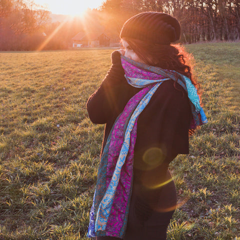 A woman is standing in the sunshine, bundled up in dark clothes and a blue and pink reclaimed sari medley scarf