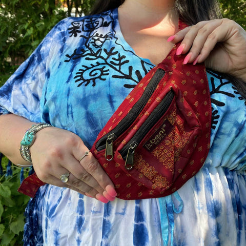 A person wearing a blue tie die kaftan is holding up a bright red and gold sari silk fanny pack.