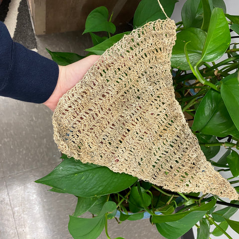 A hand is holding onto a large, triangular hammock made out of single ply hemp against a lush green leafy plant.