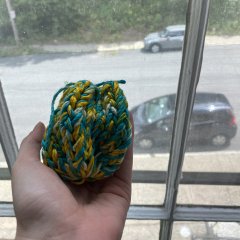 A hand is holding an unsoaked reusable water balloon, knit out of blue and yellow yarn. The person is standing at a window, staring down at the street.
