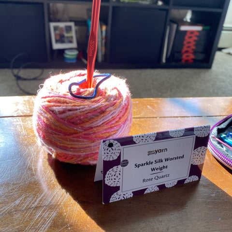 A cake of rose quarts sparkle worsted weight silk yarn in different shades of pinks is resting in the sun on a wooden table. There's a red metal crochet hook stuck into the cake and a purple yarn tag is posed in front of the cake.