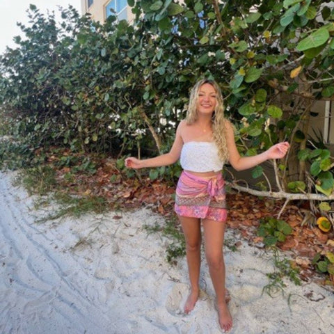 A woman with light blonde hair is standing in a sandy mangrove, wearing a white top and a pink medley scarf as a skirt.