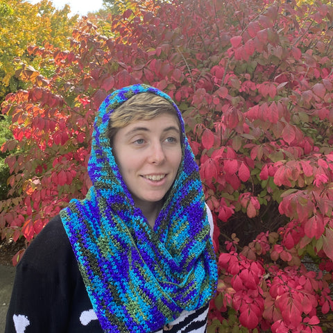 A person with short blond hair is standing beside a bright red autumnal bush, wearing the blue and green scoodie around their head and neck.