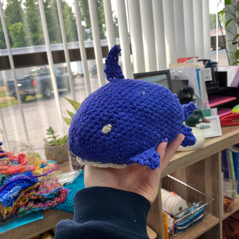 A hand is holding an amigurumi stuffed shark made out of blue and white worsted weight yarn. In the background you can see a shelving unit filled with yarn projects and books. As well as a large window with white plastic blinds.