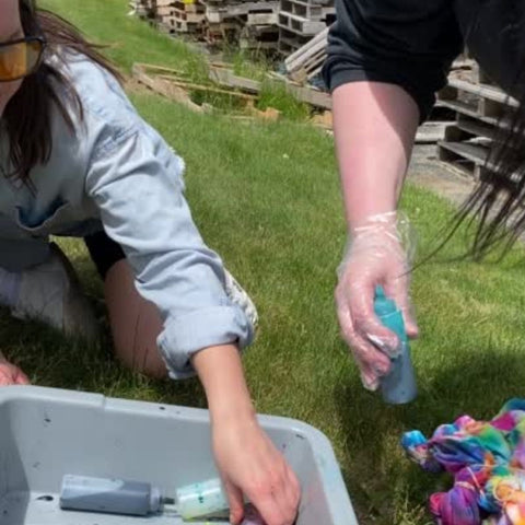 Two women are outside, reaching into a gray bin to grab bottles of dye that they will use to tie dye their white t-shirts.