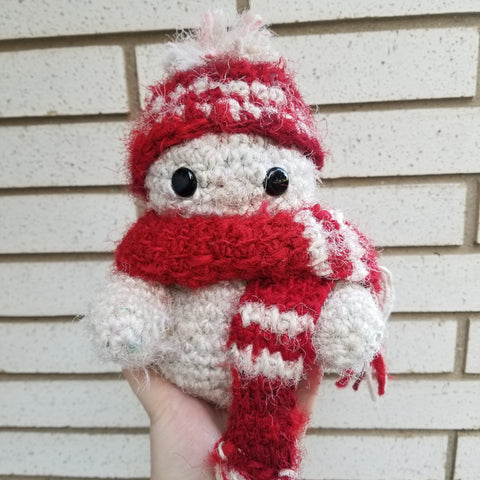 Against a white brick wall, a hand is supporting a fuzzy white snowman wearing a red and white chevron scarf and hat.