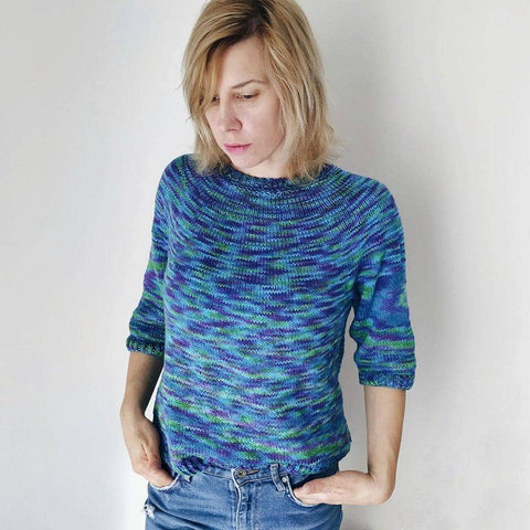 Designer wearing simple seamless sweater in colorway peacock with jeans in front of a white background.