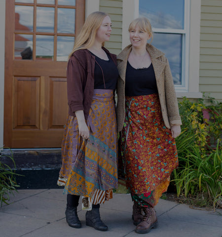 Two women are walking together, wearing open sweaters and fall-colored sari wrap recycled skirts.