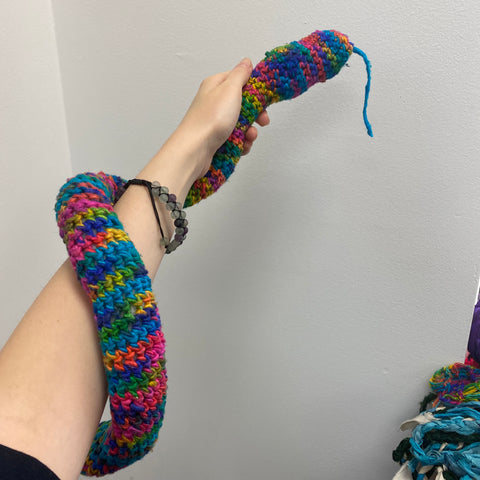Against a white wall, a pale arm is holding a rainbow amigurumi snake wrapped around their arm.