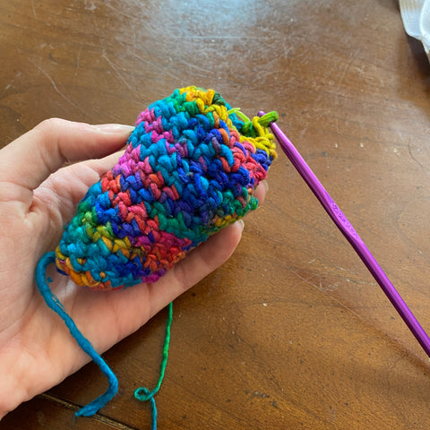 On a wooden table, a hand holds the 'head' of the amigurumi snake made out of rainbow worsted weight yarn in watercolors.