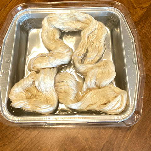 On a table, in a silver metal foil square tin, is a loose skein of white lace weight silk yarn soaking in salt water.