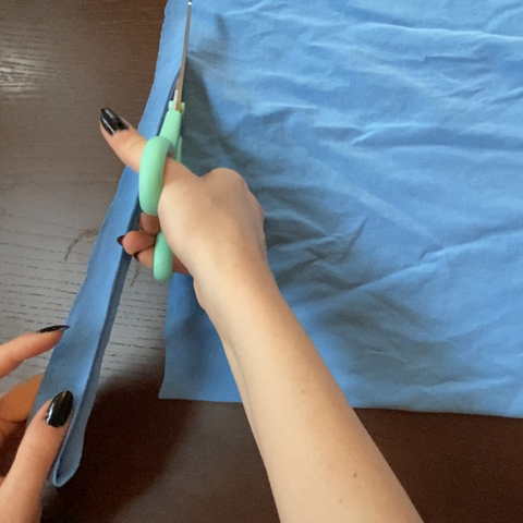 A pair of hands with black nails is using a pair of teal scissors to cut strips out of the light blue t-shirt.