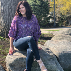 A woman with long dark hair is sitting on a rock, wearing a pair of jeans and a purple crochet top