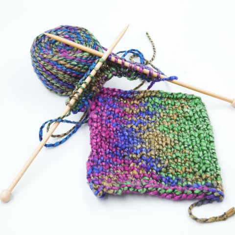A swatch of multi-colored yarn, made with the stockinette stitch, is laying on a white table. Behind the swatch is a skein of the same yarn and a pair of wooden knitting needles.