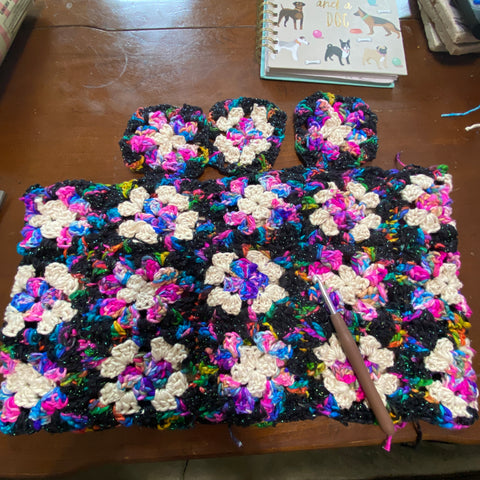 The black, white, and pink/blue granny square top is laid out on a wooden table, near the brown crochet hook used to make them, and a little journal with dogs on the cover.