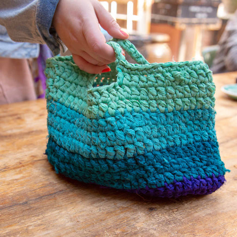 A hand is holding the handles of a blue ombre market tote bag. The bag is being set down on a wooden table.