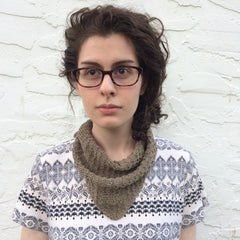 A woman wearing glasses and curly dark hair is facing the camera somberly. Around her neck is the poppyseed scarf, made out of a dark tan herbal dyed yarn.