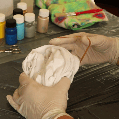 A person wearing rubber gloves is wrapping a white t-shirt up into a tight ball and then wrapping rubber bands around the shirt.