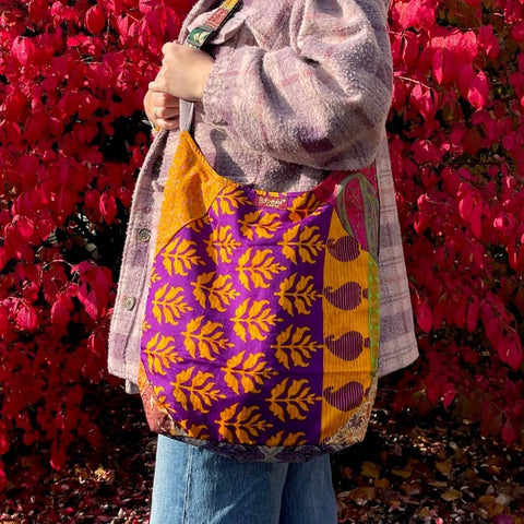 A woman wearing a pink plaid coat and a handmade sari silk bag, while standing in front of a bright red bush.