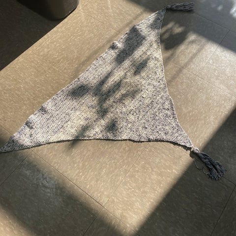 A large grey triangular scarf is laying on the ground, in the sun.