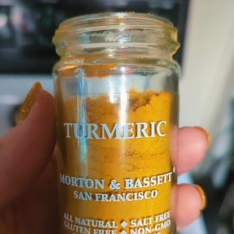 A hand with brightly colored yellow nails is holding a clear bottle of turmeric spice.