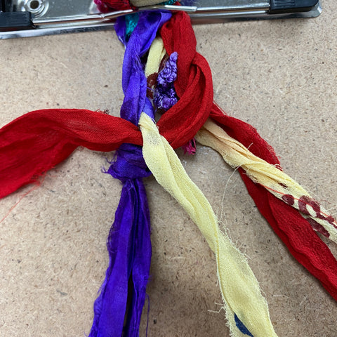 Attached to a clip board, six strands of yellow, red, and purple ribbon is being braided together