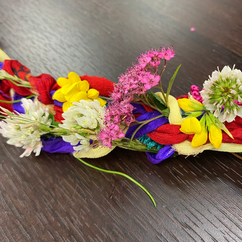 A section of the finished witches ladder, woven with red, yellow, and purple ribbon and multiple different flowers, rests on a shiny dark wooden desk.