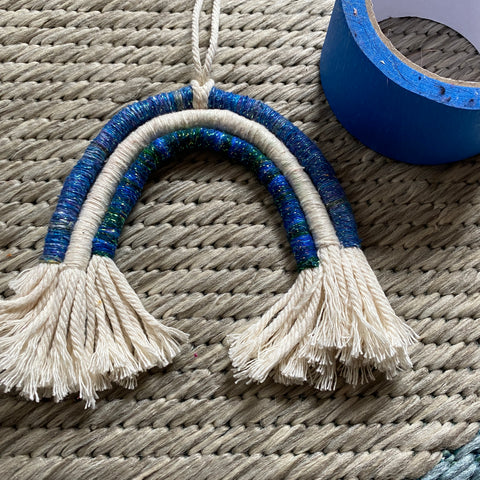 A yarn wrapped macrame rope mini rainbow made from sparkly white, blue, and green yarn is laying on a tan rug. To the right of the rainbow is a circle of blue painters tape.