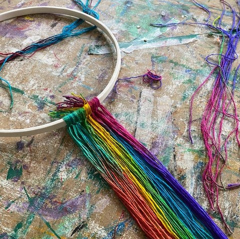 A white hoop with muticolored yarn tied to the bottom of the loop is resting against a brown artist's board. The artist's board is covered in paint marks and old painters tape.