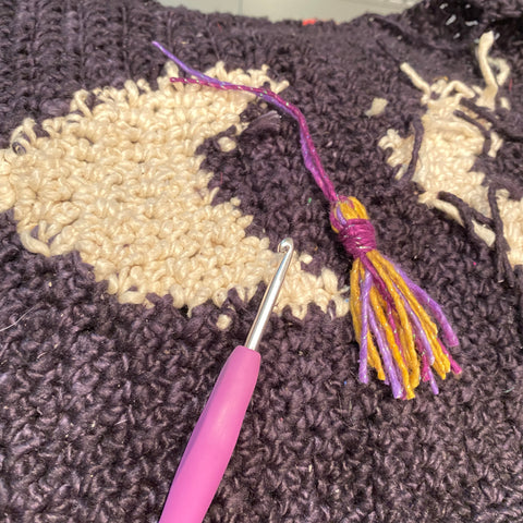 A closeup of the runner, showing a black base, a white crescent moon, a pink crochet hook, and a tassel made of purple and yellow.