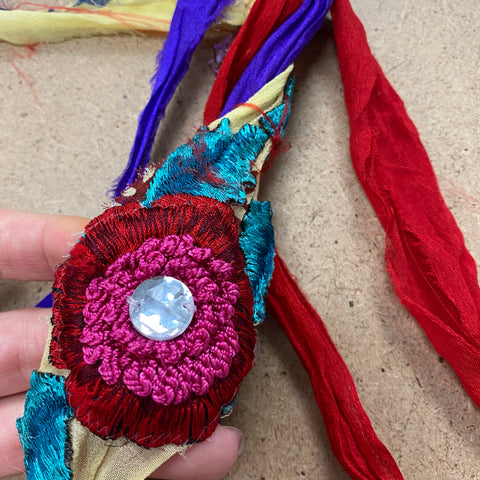 A hand holds a strand of chiffon ribbon yarn, showing an embroidered marigold made out of red, pink, and teal yarn - with a big jewel as the center of the flower!