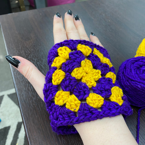 The purple and yellow granny squares have been joined together and are being worn on someone's hand.