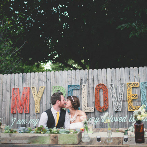 A married couple sit at their main table, kissing each other. Behind them is a large yarn sign, made of multiple colors, that reads 'My Beloved'
