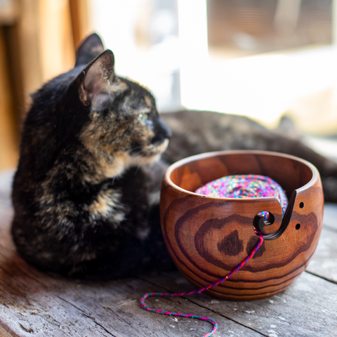 A black and brown speckled kitty laying on a wooden table, behind a wooden yarn bowl filled with handmade yarn.