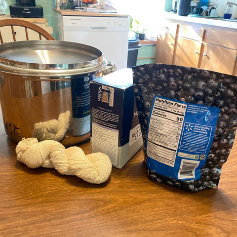 In a kitchen, on the warm wooden table sits a shiny pot, a skein of white yarn, a box of salt, and a bag of frozen blueberries