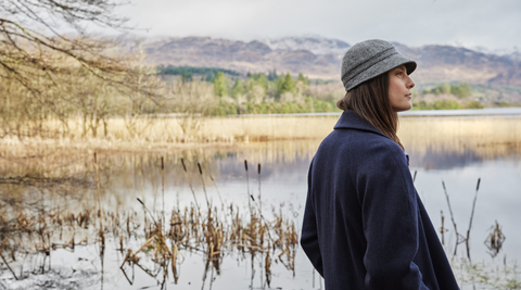Woman wearing a tweed coat and 1920's style grey tweed hat standing at a lake edge taking in the scenery