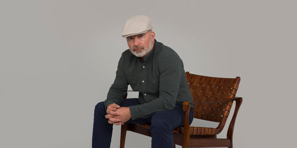 Mann sitzt auf einem braunen Stuhl und trägt eine helle Schiebermütze