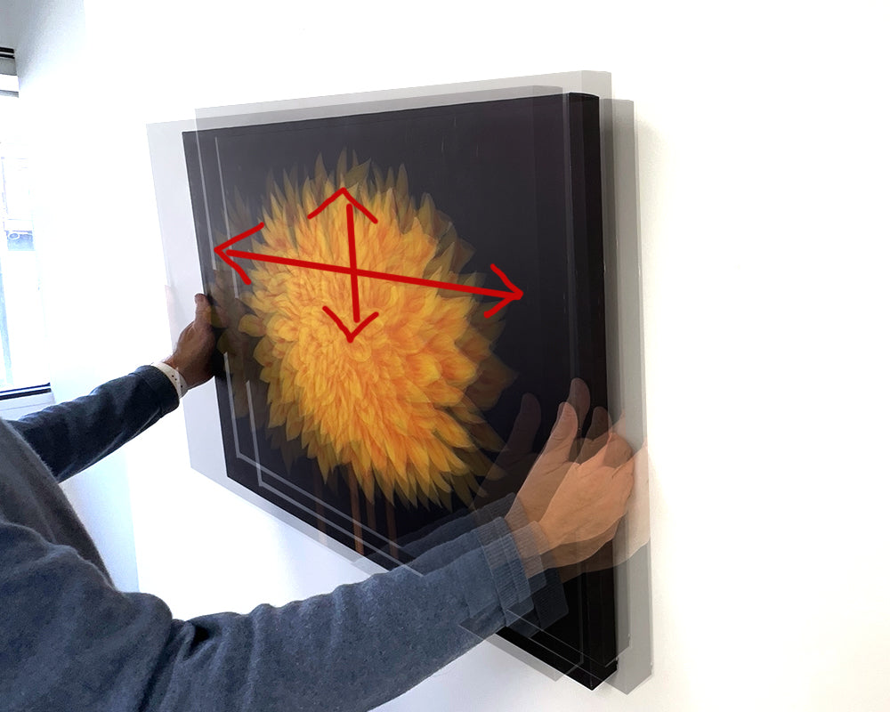 A person holding canvas art up on the wall and arrows showing it can be moved up, down or sideways to adjust the location because of picture wire installed to hang it.