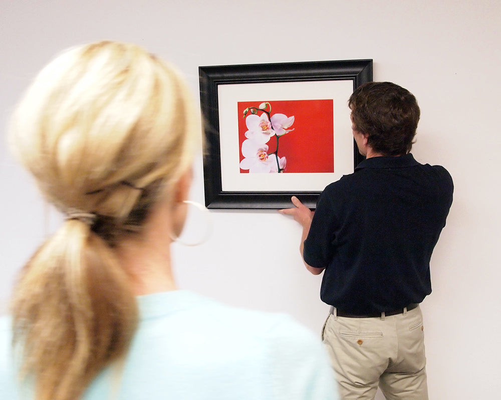 couple hanging a picture installed with two keyhole hangers: one person holds the picture while the other stands back to check the wall position