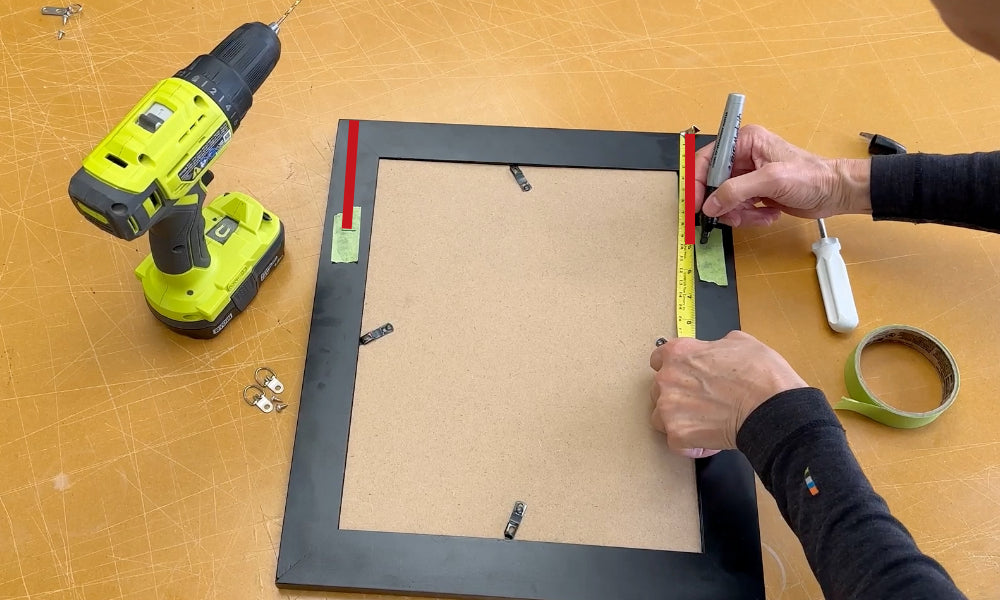 Back of a picture frame on a work table and a person marking the same distance from the top of the frame on both sides of the frame to show where to position the D-ring hangers.