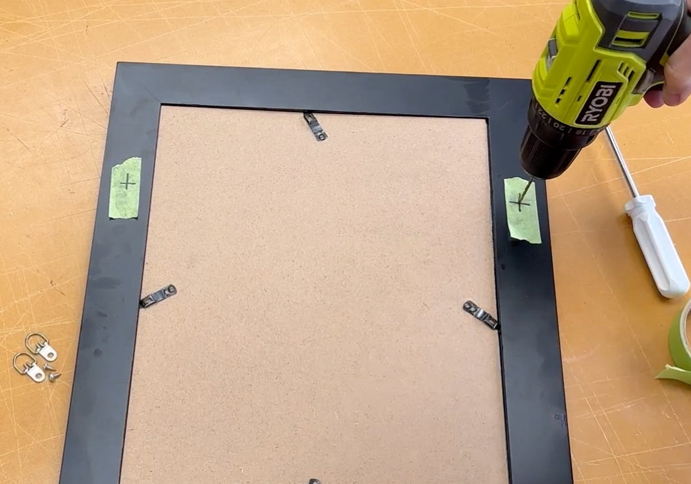 Image of a person drilling starter holes in the back of a picture frame on the marks previously made on the frame