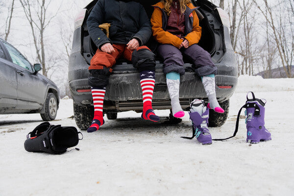 A couple seated on tailgate wearing Captain Stripe and Yeti ski socks, about to pull on ski boots