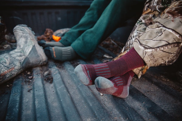 A man's feet and a woman's feet, both wearing darn tough wool hunting socks, resting on tailgate