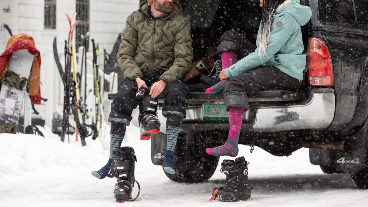 A skier and a snowboarder seated on tailgate comparing socks from darn tough