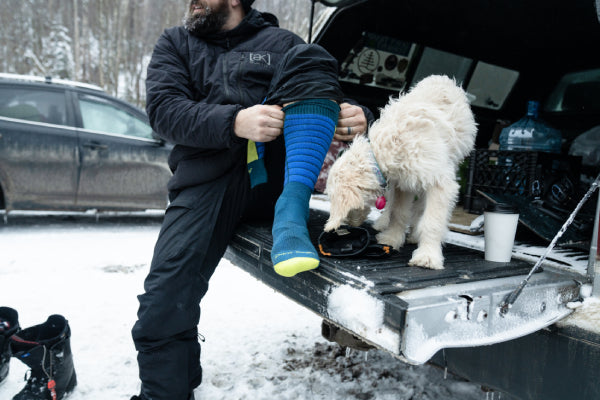 Man putting on Darn Tough Ski Socks while in trunk with a dog