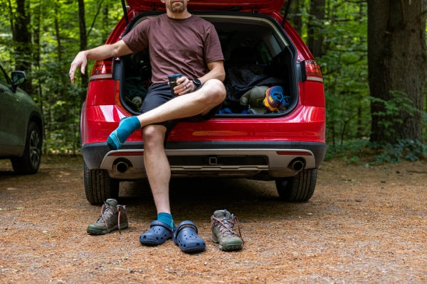 Man sitting on the back of a red car taking off boots and putting on crocs