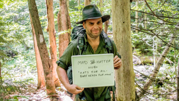 Mad Hatter wearing his hat with a sign that says "hats for all, bare heads for none"